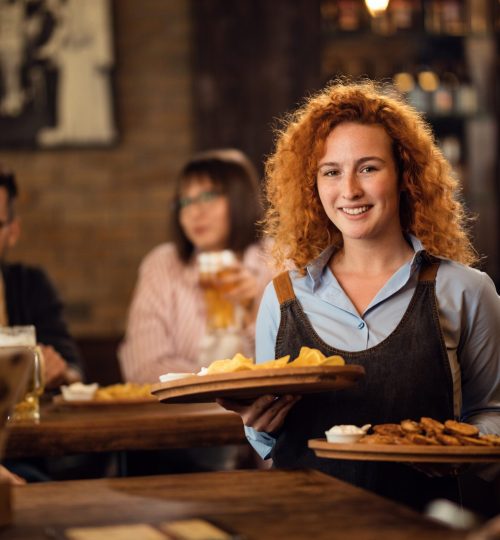 Happy redhead waitress serving customers in a restaurant and bringing food at their table.